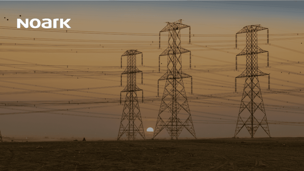 Tres torres de alta tensión se alzan en un paisaje árido al atardecer, con numerosas líneas eléctricas y dispositivos de circuitos de control integrándose sutilmente en el cielo.