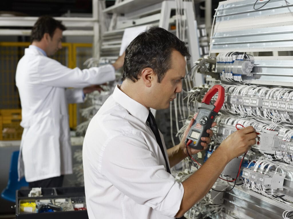 Dos técnicos trabajan en cuadros eléctricos; uno utiliza un multímetro, mientras que el otro inspecciona el cableado. Están en un laboratorio y llevan camisas blancas.