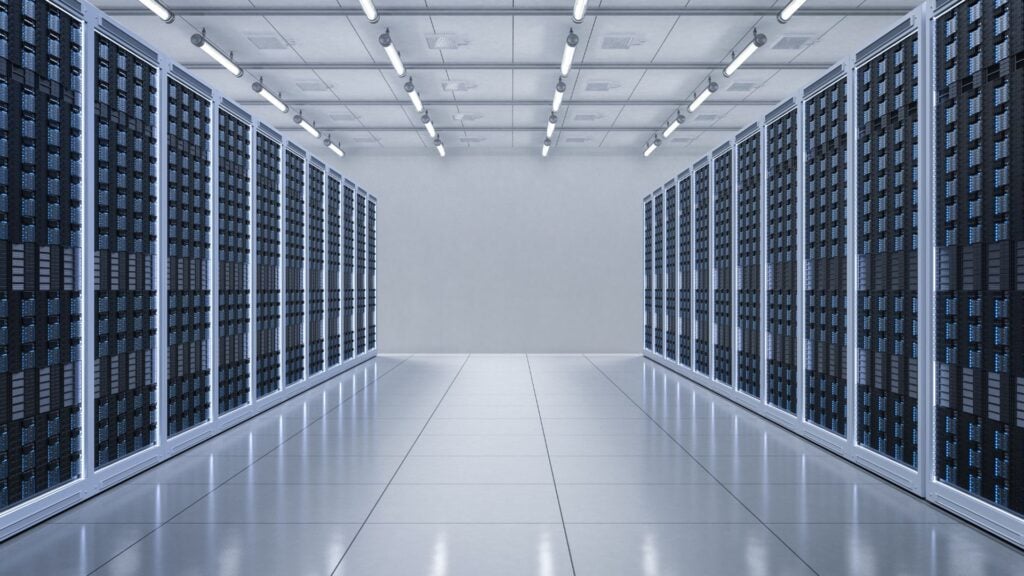 Un moderno centro de datos con hileras de bastidores de servidores, optimizados para el almacenamiento digital, se alinean a ambos lados de un pulido pasillo bajo las brillantes luces del techo.