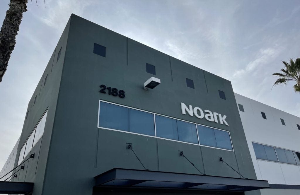 Edificio de dos plantas con el logotipo "Noark" y el número 2188 en la fachada. La estructura tiene un diseño moderno con grandes ventanales y un exterior verde azulado. Cielo y palmera parcialmente visibles.