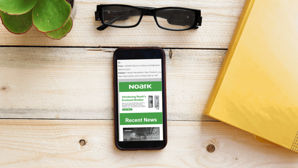 Un smartphone electrifica la escena al mostrar un correo electrónico de NOARK sobre una mesa de madera, rodeado de gafas, una planta y una carpeta amarilla: un curioso buzón de la comunicación moderna.