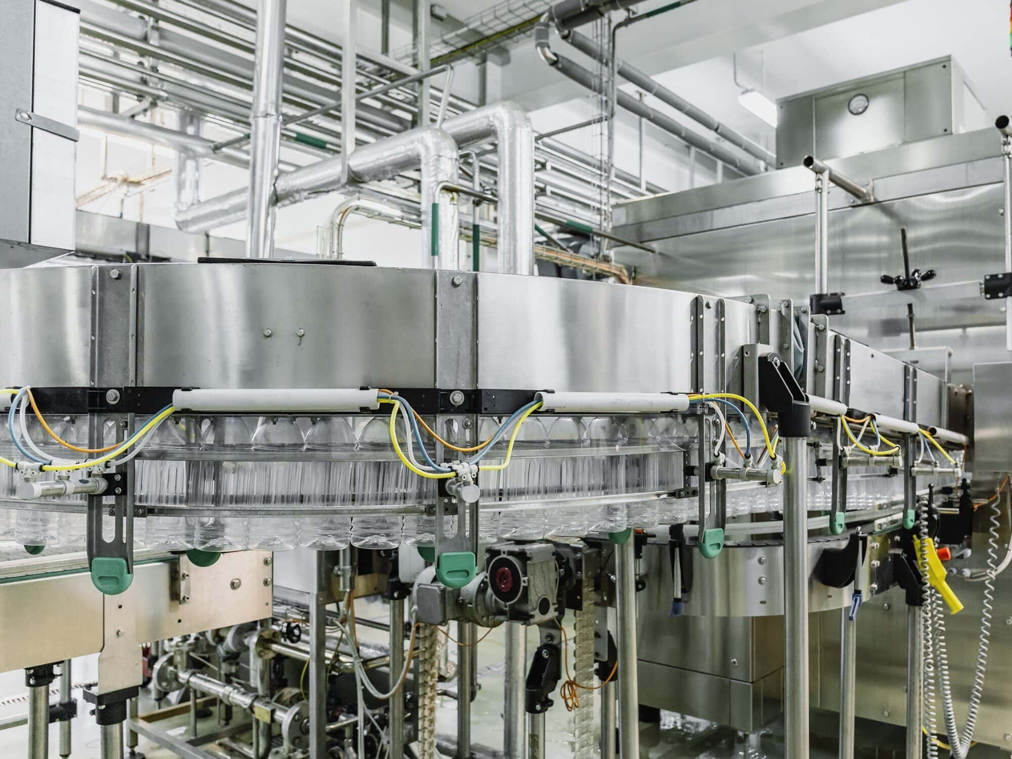 Una moderna línea de embotellado automatizada en una fábrica, con filas de botellas transparentes sobre una cinta transportadora en medio de una red de tuberías y maquinaria.