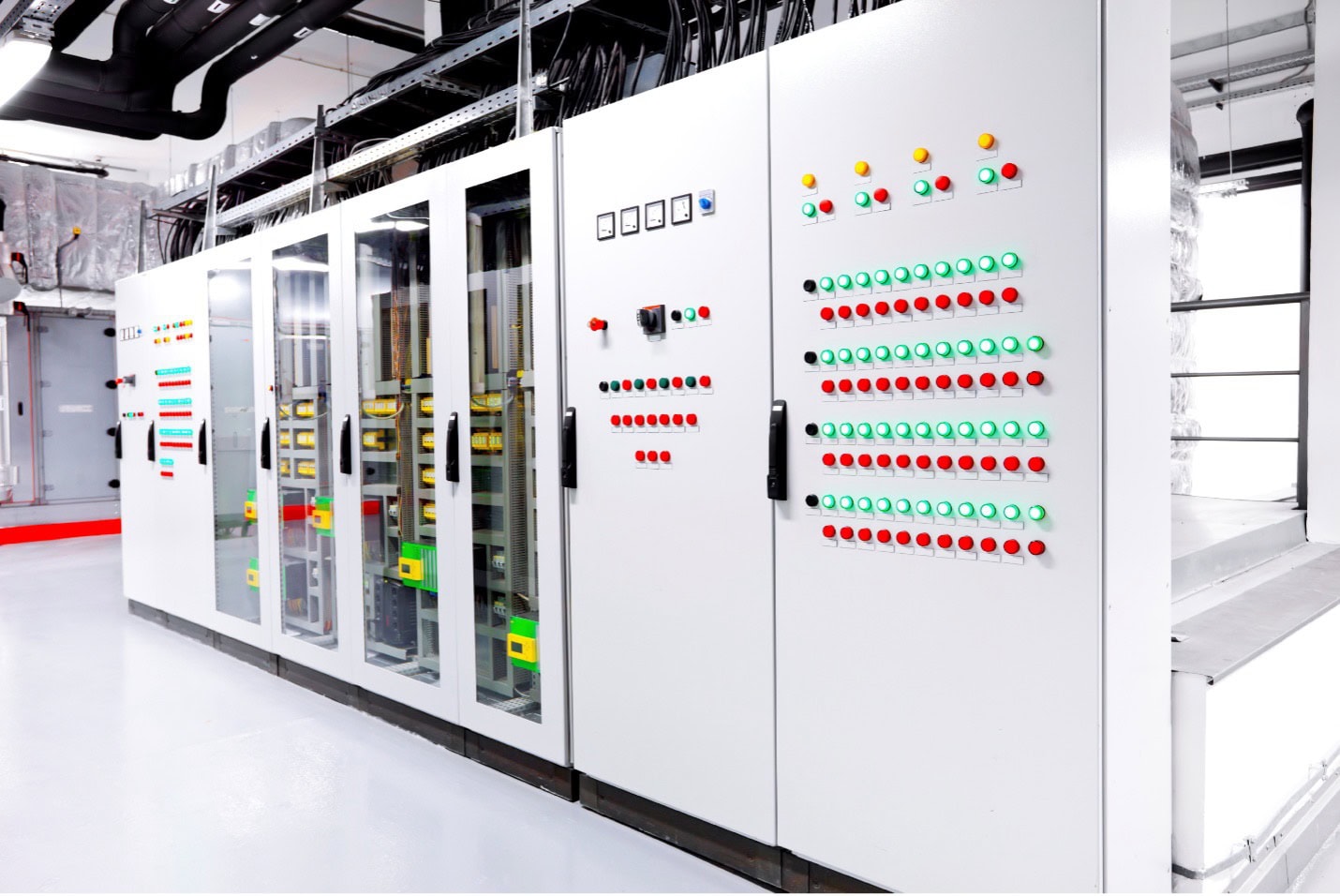 Una hilera de paneles de control eléctrico blancos con botones de varios colores, interruptores y pequeñas pantallas en un entorno industrial muy iluminado.