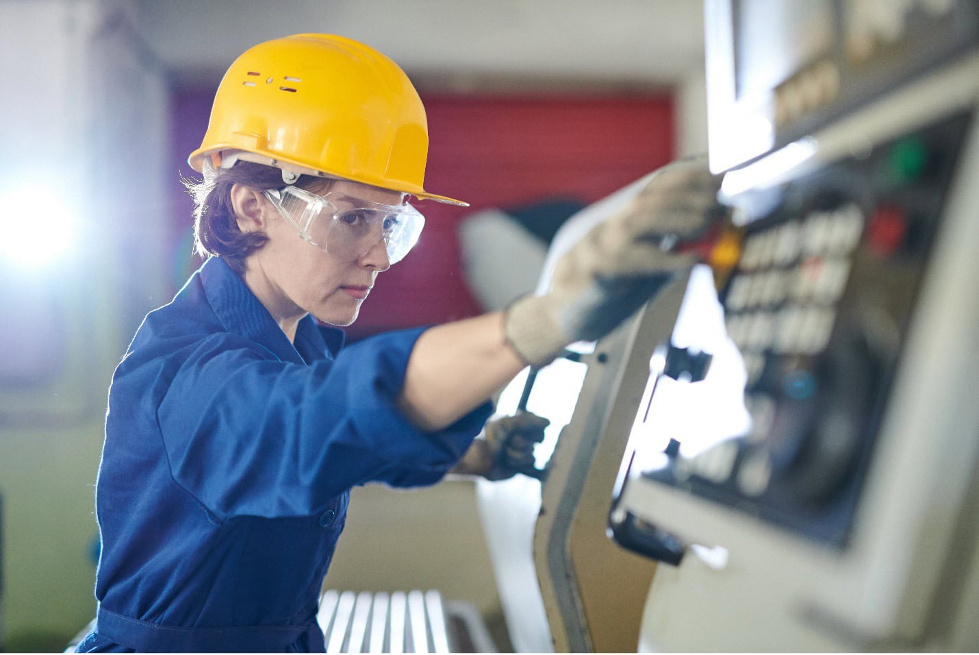 Una persona con casco amarillo y gafas de seguridad maneja un panel de control en un entorno industrial. Lleva atuendo de trabajo y guantes azules.