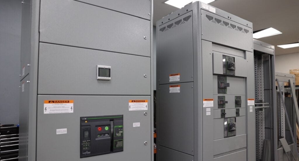 Dos grandes armarios eléctricos grises, parecidos a cuadros industriales, presentan paneles de control y etiquetas de precaución en una sala bien iluminada.